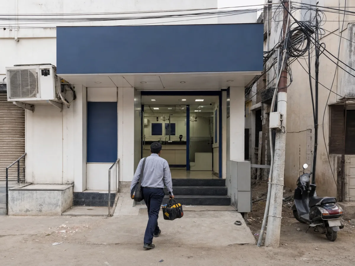 A field engineer carrying a tool bag walks up to a small bank branch on an Indian street